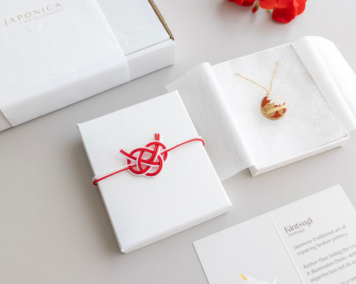 A styled flat-lay of a Japanese pendant necklace on a gold chain, displayed inside an open white presentation box. The necklace features a gold heart pendant with a red and white marbled pattern.